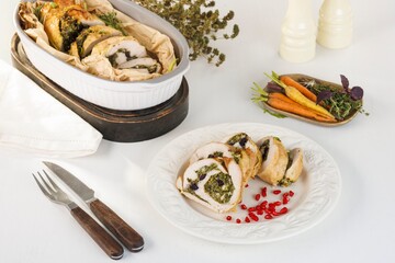 Sliced turkey breast roll ups are arranged on a white plate with pomegranate seeds. Baked carrots with herbs are presented on a wooden plate. All sitting on a white table