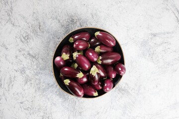 A collection of small, shiny purple eggplants fills a dark bowl. The bowl sits on a light grey surface with a textured, mottled appearance creating a simple, yet elegant still life