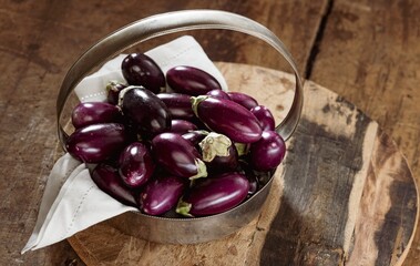 A Collection of small, shiny purple eggplants are Piled in a vintage metal basket. The basket sits on a textured wooden chopping board next to a white cloth