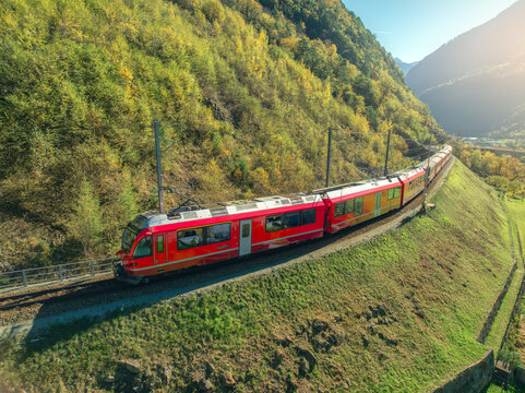Aerial view of modern red train in alpine mountains, green meadows at sunset in autumn. Bernina Express, Switzerland. Top view of train in Alps, winding railroad in fall. Railway station. Travel