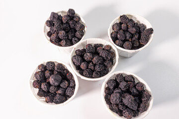 Top view of five white paper cups filled with dark, shriveled, dried chokeberry berries and arranged on a white surface.