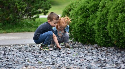 Little boy and red-haired toddler girl crouching together, exploring gravel stones in park. Children learning, curiosity, friendship, happy outdoor play, and childhood discovery moments.