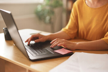 Naklejka premium Individual working on a laptop at a wooden table, with a coffee cup and papers scattered nearby, in a bright and inviting indoor environment while natural light filters through.