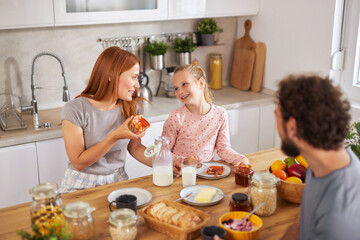 A mother and daughter share a joyful breakfast moment in a well-lit kitchen. They smile while enjoying food, surrounded by fresh ingredients and breakfast items on the table.