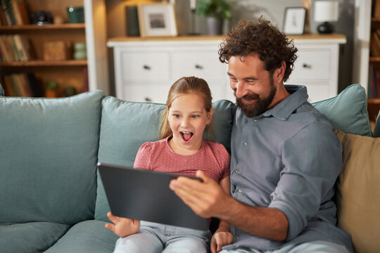 A man and a young girl share a joyful moment as they watch a tablet together. The living room is filled with warm light, creating a cheerful atmosphere for their bonding time. - Powered by Adobe