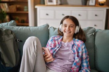 Obraz premium A cheerful teenage girl is seated on a couch, wearing headphones and smiling. She listens to music while holding a phone, dressed in a comfortable outfit, enjoying her free time at home.