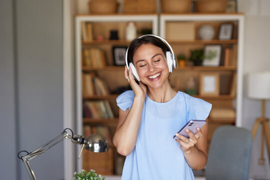 A woman stands in a well-decorated home office, smiling as she listens to music through headphones while holding a smartphone, creating a lively and productive atmosphere.