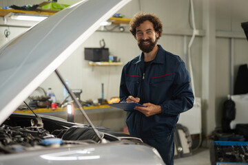 Man wearing mechanic uniform works on a car in a repair shop. He smiles while taking notes on a clipboard, surrounded by tools and equipment, indicating a busy and productive environment.