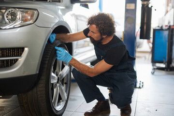 A car mechanic inspects a tire closely while kneeling beside a vehicle in a well-lit repair shop. The professional wears blue gloves and is attentive to detail, ensuring quality work.