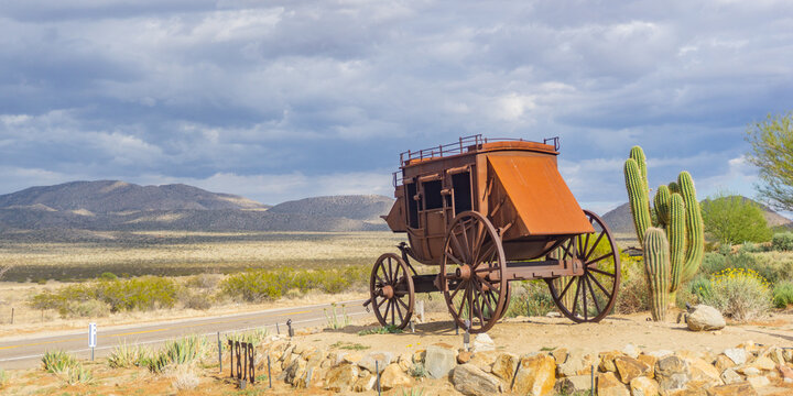 Old stagecoach sits on a rocky outcrop overlooking a desert landscape under a cloudy sky backdrop
