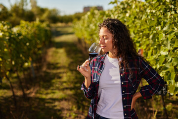 A woman enjoys a glass of red wine while standing in a vineyard. Rows of grapevines surround her under the warm sunlight, highlighting the family's dedication to wine production.