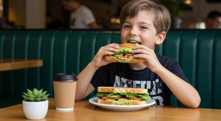 A young boy eats a sandwich at a restaurant table, with another sandwich and a coffee cup nearby. The background features a green booth and blurred figures.