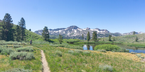 Naklejka premium A scenic view of a mountain range with snow, trees, a lake, and a hiking trail under a clear blue sky