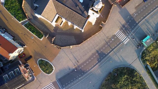 Aerial view of a public square with red‑tiled roof buildings, a central triangular‑roof pavilion, lounge chairs, sports court markings, and landscaped areas casting long afternoon shadows.