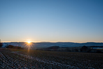 Sunrise above distant winter hills with snowy farmland in foreground, cold rural morning landscape.