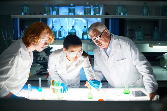 Multi-ethnic group of medical scientists examining substance in test tube. They bending over table with laboratory glassware and looking with concentration at experiment results