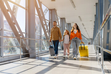 Fototapeta premium Happy european family walking side by side at airport with luggage suitcases, holding hands and smiling in sunlight, symbolizing cheerful bonding, lifestyle and holiday travel joy