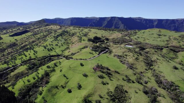 Wide drone footage over Plaine des Cafres on Reunion Island. Green plains, a scenic valley, scattered trees, and a peaceful road with passing cars create a tranquil mountain landscape.