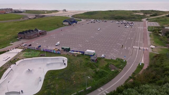 Aerial view of coastal area with skatepark, large parking lot, event gathering, buildings, and ocean backdrop blending recreation, infrastructure, and natural scenery.