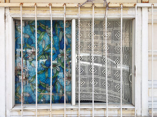 the left with vibrant floral stained glass, the right with a sheer crochet curtain. Rusty metal bars and a weathered wall frame the window, blending bright patterns with aged, neglected textures.