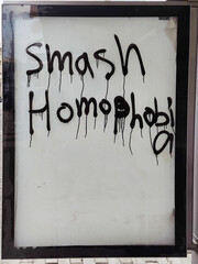 A message painted on glass outdoors reads “smash homofobia” in black paint with drips.