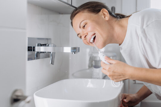 A young woman uses a water flosser to clean her teeth in the bathroom in the morning. Concept: morning routine and oral hygiene at home.
