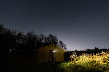 A solitary person with a headlamp stands in the doorway of a remote wooden cabin under a vast...