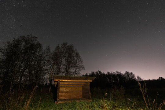 A rustic wooden log shelter or lean-to sits in a quiet meadow under a brilliant, star-filled night sky.