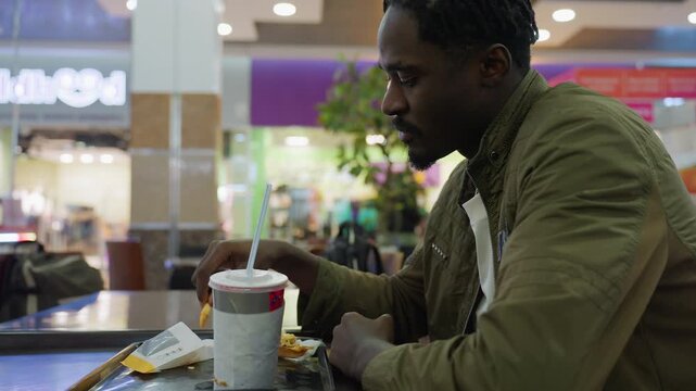 Side view of young man in olive jacket sitting alone at food court table eating fries, with paper cup and empty food pack on tray in front of him surrounded by blurry mall background