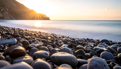 A tranquil scene of a pebbled beach at sunrise. Soft light bathes the smooth stones, gentle waves and a hazy horizon. Sunbeams burst through the sky
