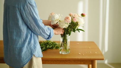 Woman arranging peony and rose flowers in a glass vase, creating a beautiful bouquet for home decoration, enjoying a peaceful floristry hobby activity in soft daylight