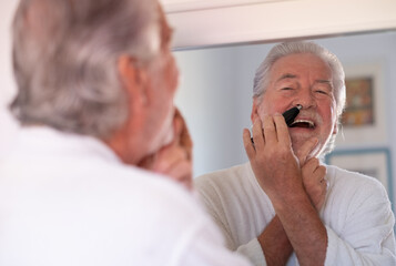 Portrait of senior bearded man in bathrobe while remove nose ear hair with help of little comfy trimmer looking at himself in the mirror