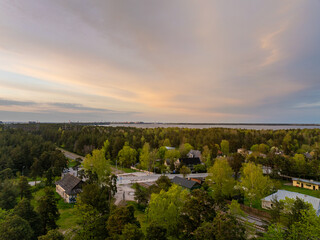 Golden-hour aerial view of Manniku district, Tallinn, Estonia, showing pine forest, scattered houses, railway tracks and Lake Ulemiste under soft pastel evening sky (Estonian - Manniku, Ülemiste)