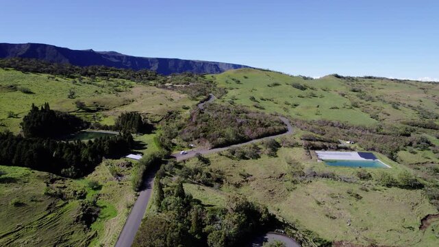 Drone footage following a sinuous road at Plaine des Cafres on Reunion Island. Cars traverse the scenic route, leading to a dramatic, vertiginous cliff and valley view.