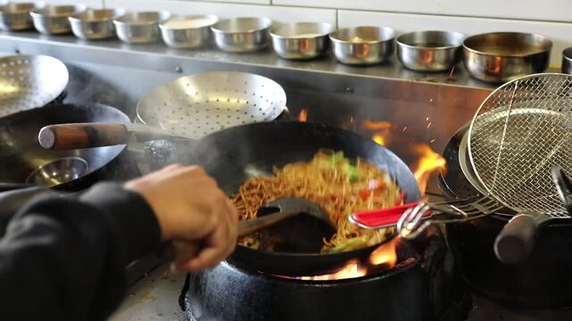 Chef stir frying Asian noodles with vegetables in hot wok at restaurant kitchen, with steam and flame.