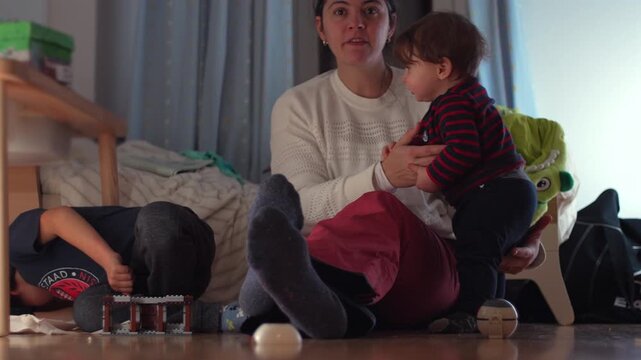 Mother supporting toddler to keep balance while older child plays beside them on bedroom floor, intimate family bonding moment of parenting, care, and learning at home