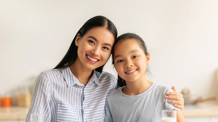 Young asian mother and daughter enjoying fresh homemade cookies, drinking milk and smiling to camera while sitting at table in kitchen, spending time together at home