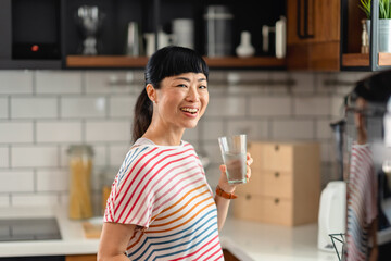 Smiling Japanese woman holding a glass of water in modern kitchen. Cheerful Asian woman promoting hydration, wellness and healthy lifestyle with natural light and relaxed mood.
