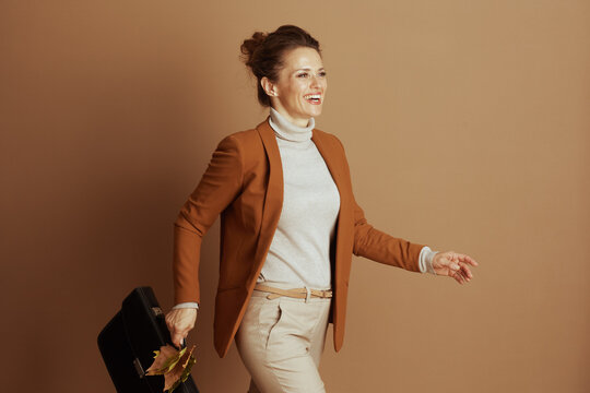 A cheerful businesswoman moves confidently, holding a briefcase decorated with autumn leaves. Dressed in a stylish blazer and turtleneck, she embodies professional success and dynamic fall fashion.