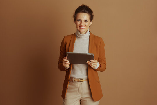 A confident, smiling woman in smart casual business attire holds digital tablet horizontally, looking at the camera. Her brown blazer and beige turtleneck convey professional yet approachable demeanor