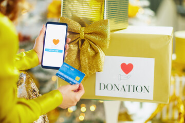 A close-up shows hands holding smartphone displaying donation app and credit card over gold gift box. The festive background of blurred lights highlights theme of charitable giving during holidays