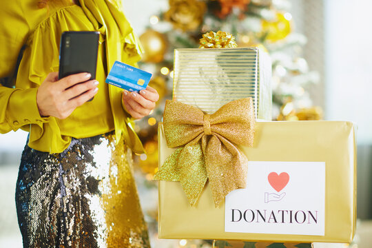 A woman in yellow blouse and sequined skirt holds smartphone and credit card, ready for holiday donation. Gift boxes and blurred Christmas tree create festive backdrop, emphasizing generosity