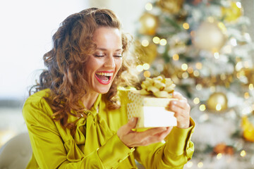 A joyful woman with curly brown hair, wearing a bright yellow blouse, laughs with eyes closed while holding a golden Christmas gift box. A softly blurred festive tree shines behind her.