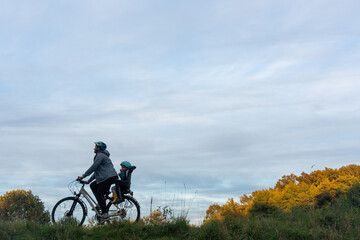 Obraz premium Mother cycling with child in bike seat along countryside path under dramatic cloudy autumn sky with golden trees and copy space for text. Concept of family outdoor activities and eco-friendly travel.