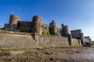 Conwy Castle with round defensive towers and fortified walls rising above the estuary at low tide under a clear blue sky © Philip