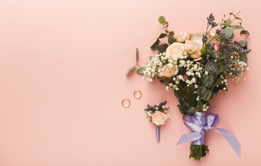 A beautiful wedding bouquet made of soft pink roses, greenery, and a delicate blue ribbon is placed next to two golden rings and a matching boutonniere on a pastel pink surface.