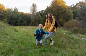 Obraz premium Joyful mother and laughing child running hand in hand through autumn meadow with golden trees. Concept of family bonding and healthy outdoor lifestyle with children.
