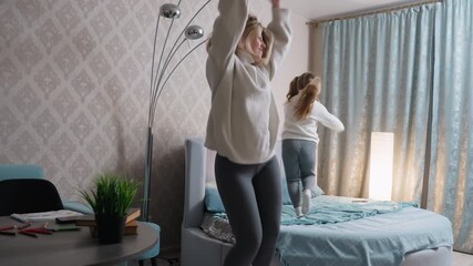 Mother and daughter wearing white sweaters and gray leggings joyfully jump on bed in cozy bedroom, enjoying playful family bonding, fun activity, carefree laughter, happiness, energy, and childhood