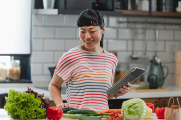 Smiling Japanese woman standing in kitchen with digital tablet and fresh vegetables. Cheerful Asian woman enjoying cooking at home, representing healthy lifestyle and happiness