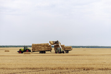 Obraz premium Farm Machinery at Work: A Tractor with a Trailer Loading Hay Bales Under Cloudy Skies in a Vast Golden Field of Straw